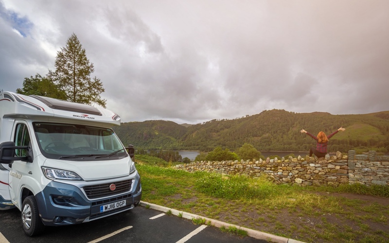 Luxury motorhome parked by the side of a lake, with girl sat on a wall admiring the view in the lake district. 